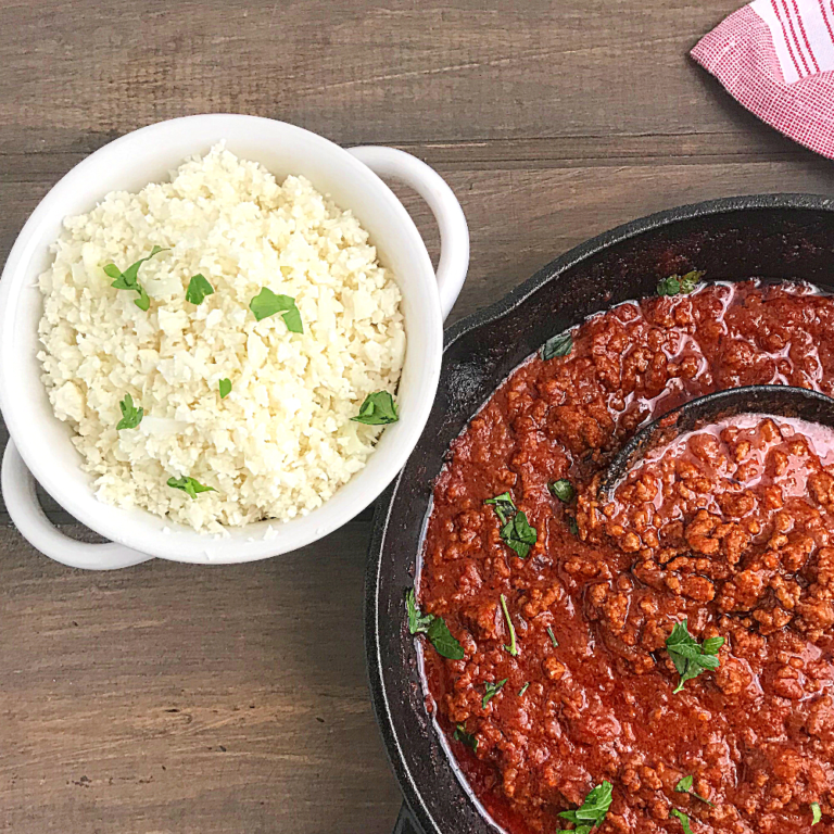 cauliflower rice in a white bowl with a skillet of tomato sauce