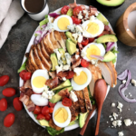 a white plate of cobb salad, showing slices of boiled eggs, chicken breast, cherry tomatoes, crumbled blue chese and onions
